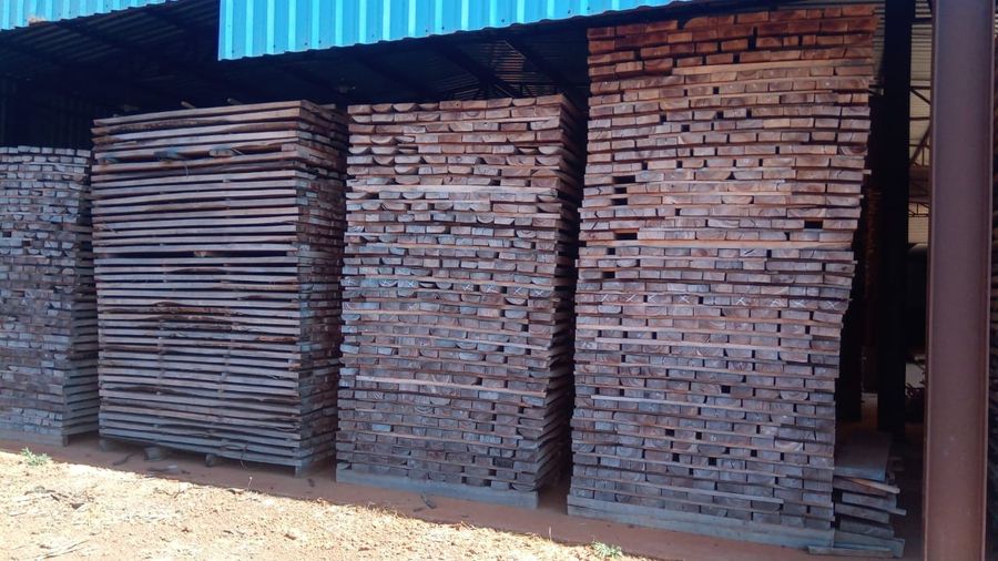 Pátio externo da madeireira com fardos de tábuas escuras sob o telhado azul