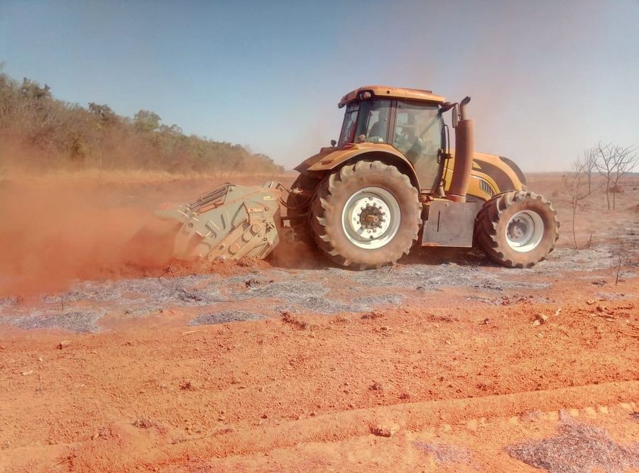 Trator amarelo com implemento levantando poeira vermelha no campo seco